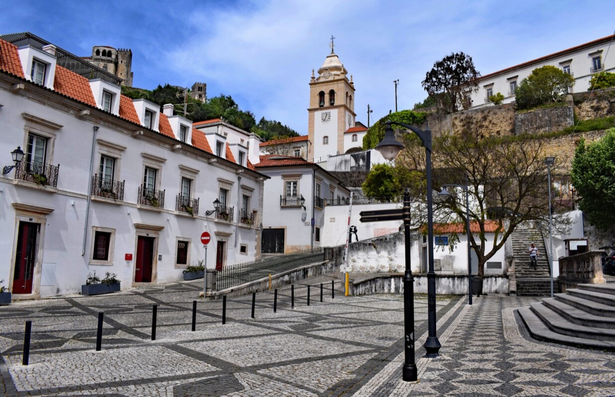Uma rua da cidade de Leiria, onde estão alguns edifícios e a torre da igreja ao fundo, num dia limpo.