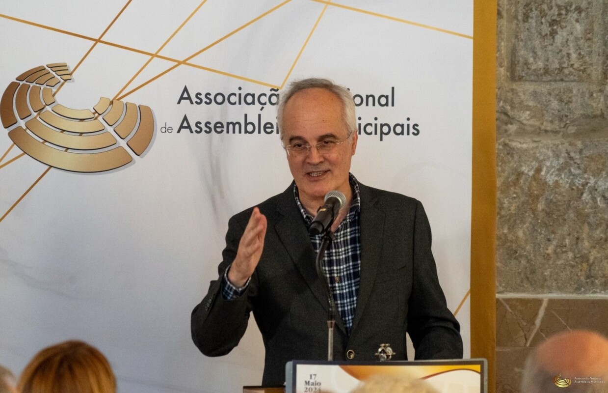 Albino Almeida, homem calvo de cabelo branco, vestido com um fato e camisa aos quadrados a discursar num palanque com microfone. Atrás, um placar com o logótipo de 'Associação Nacional de Assembleias Municipais'.