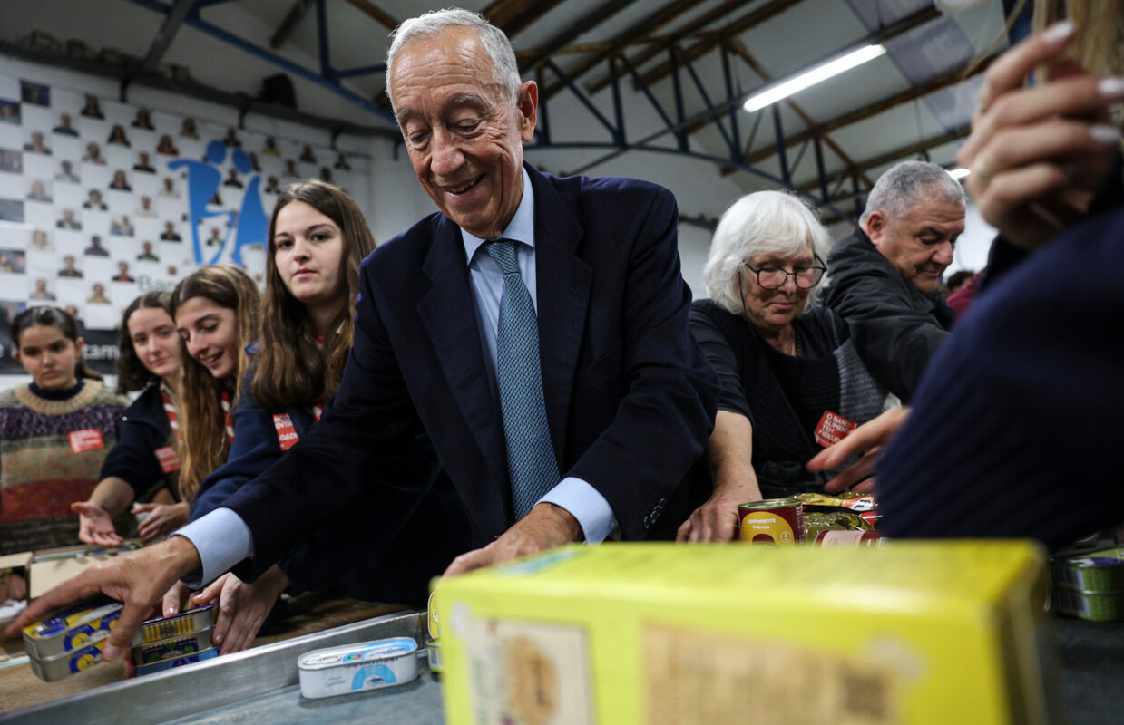 Marcelo Rebelo de Sousa, homem idoso, ao centro, com pessoas de várias idades em torno dele, com as mãos em produtos alimentares na sede de recolha do Banco Alimentar.