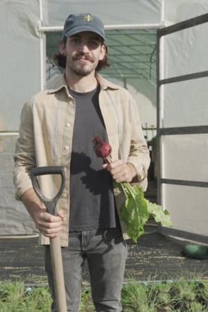 Tiago Fonteboa, fundador do projeto "Formiga Gloriosa" a segurar num engaço com a mão direita e com um rabanete na mão esquerda, com o terreno cultivado em pano de fundo